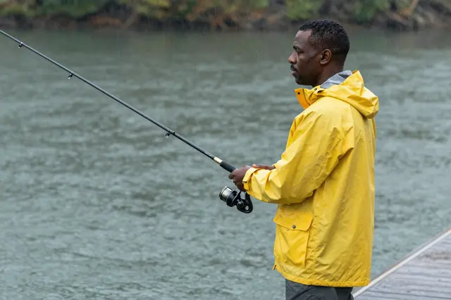 A Black man in a bright yellow rain jacket fishes from a wooden dock in light falling rain over a rippled lake.