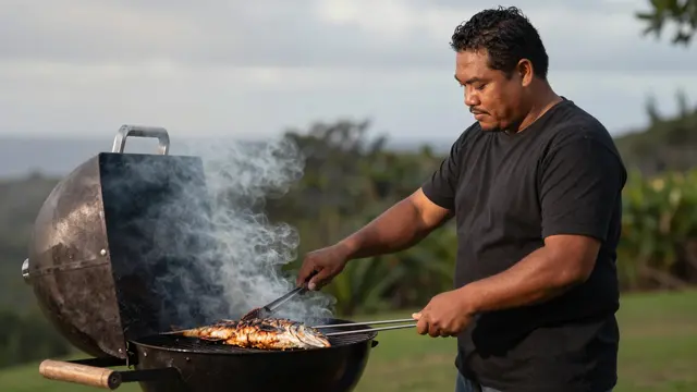 A Pacific Islander man in his thirties grills whole fish on a charcoal grill in a grassy backyard at dusk with drifting