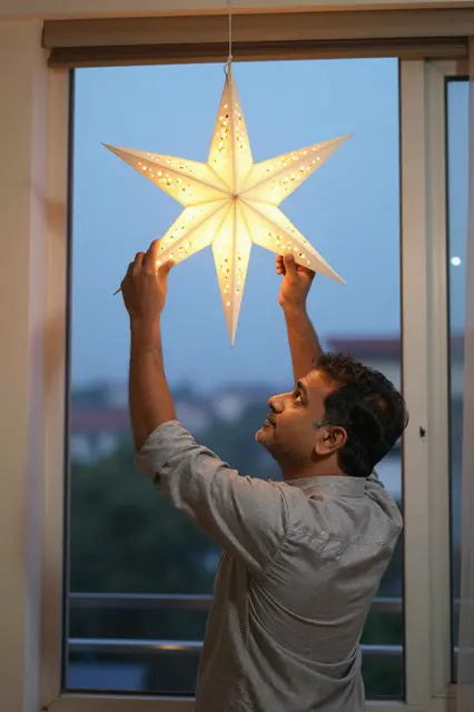 A South Asian man adjusts a glowing paper star lantern while standing by a window at dusk