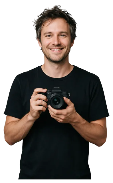 A relaxed man in a black t-shirt holds a camera at chest height and smiles at the viewer.