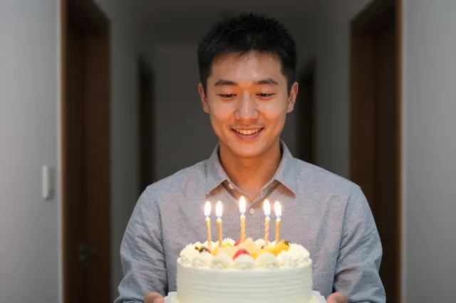 East Asian man in his thirties holds a fruit-topped birthday cake with glowing candles in a dim home hallway