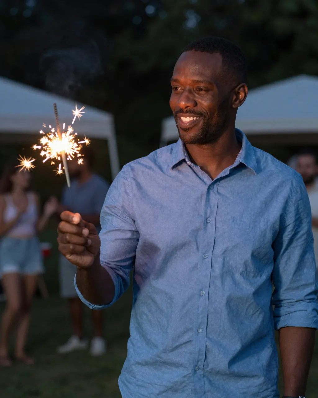 Smiling Black man in a blue shirt holds a lit sparkler at a nighttime outdoor holiday gathering