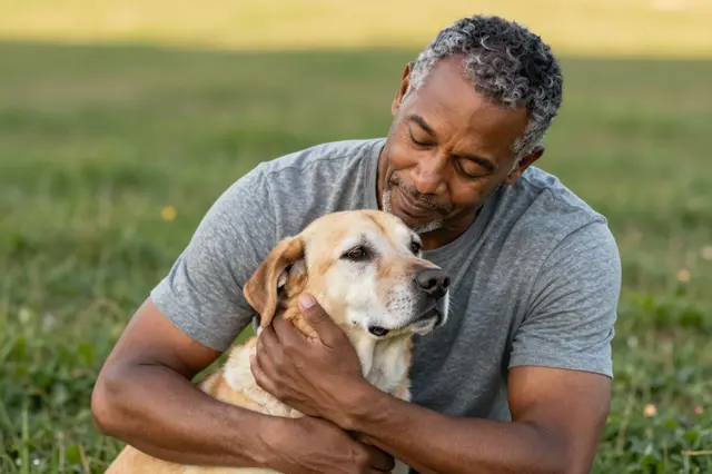 A mixed-race man in his fifties with closed eyes gently hugs an older yellow dog on grass in golden hour light