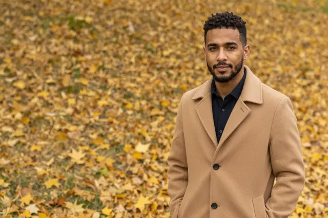 A mixed-race man in a camel overcoat stands among golden fallen autumn leaves, with copy space to his left.