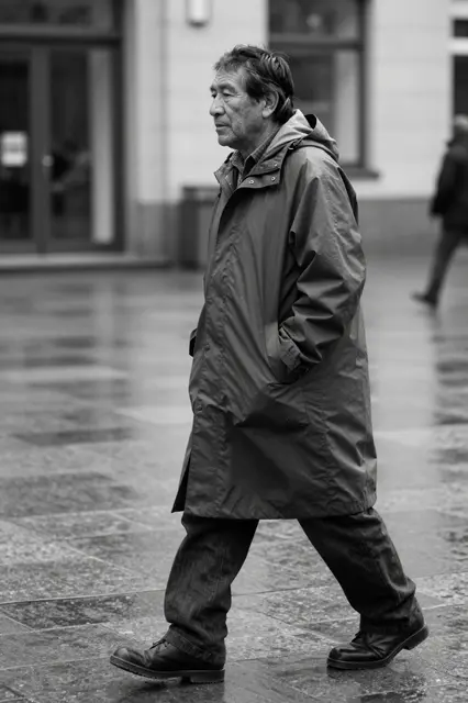 Black and white street photo of an older man in a long raincoat walking across a wet tiled plaza.