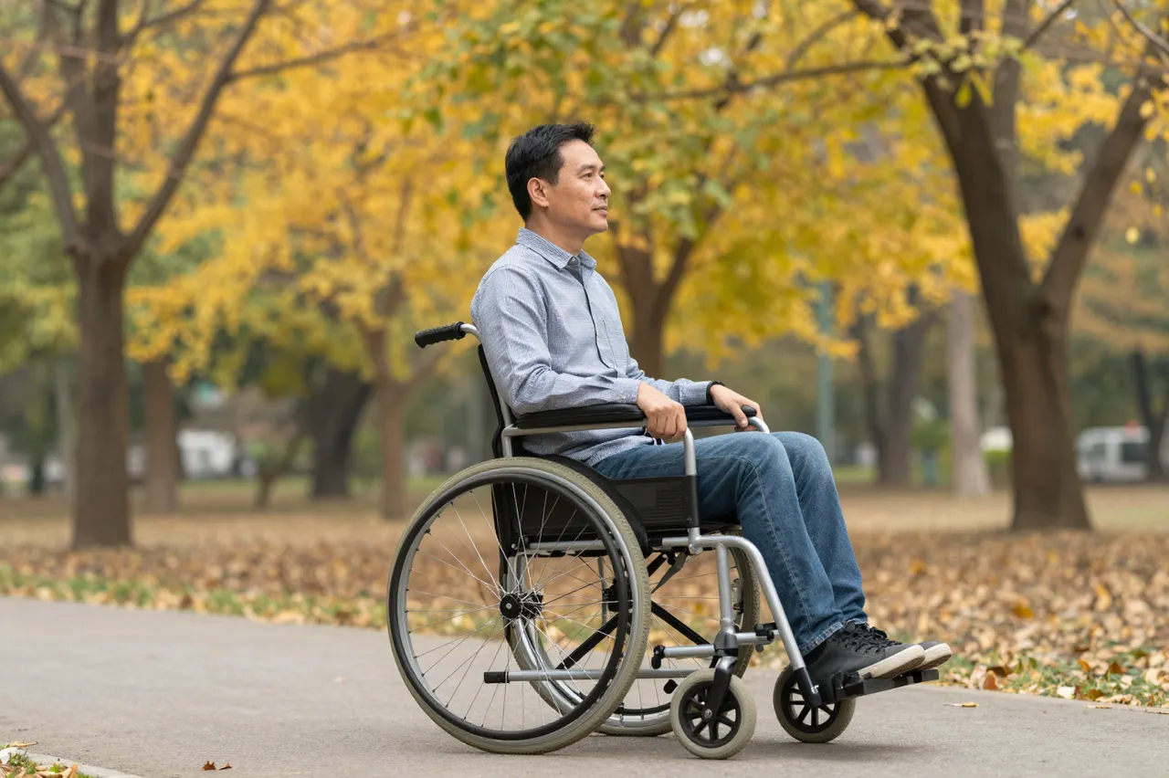 Side view of a middle aged Southeast Asian man in a wheelchair on a paved park path beneath golden autumn trees