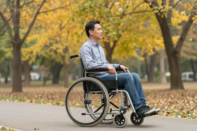 Side view of a middle aged Southeast Asian man in a wheelchair on a paved park path beneath golden autumn trees