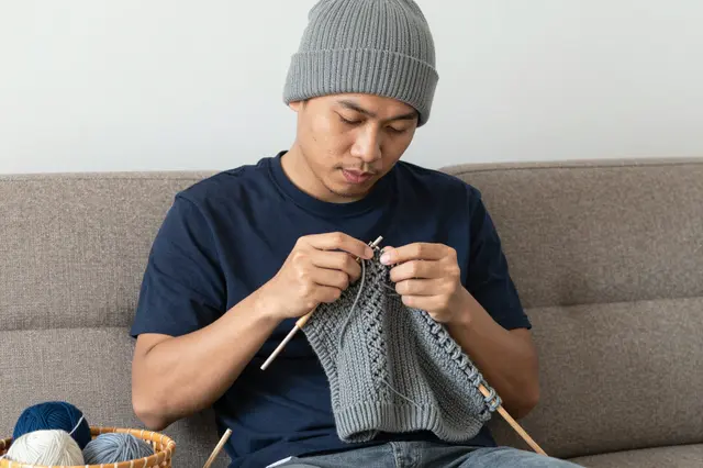 A Southeast Asian man in his thirties knits gray fabric while seated on a brown sofa, with a yarn basket beside him.
