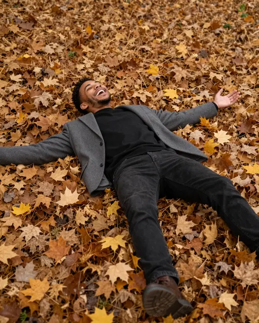 A laughing mixed-race man lies back with arms spread across a thick pile of fallen autumn maple leaves