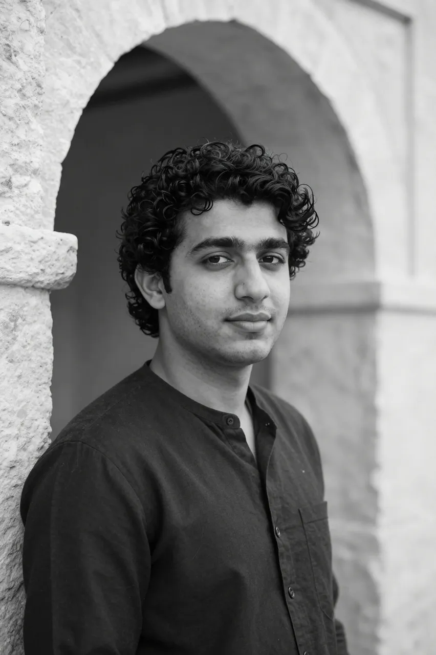 Black and white portrait of a curly haired man in his late twenties leaning against a stone archway