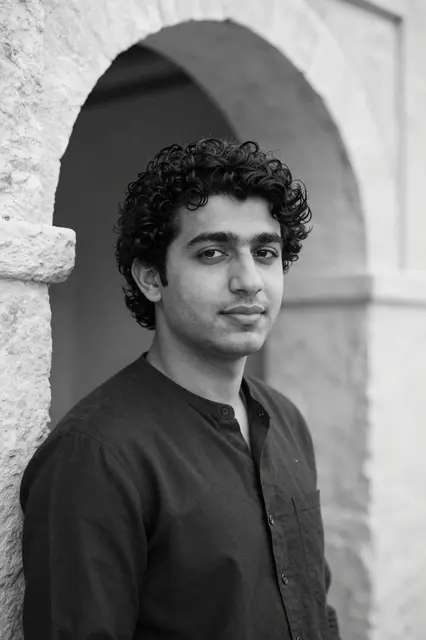 Black and white portrait of a curly haired man in his late twenties leaning against a stone archway