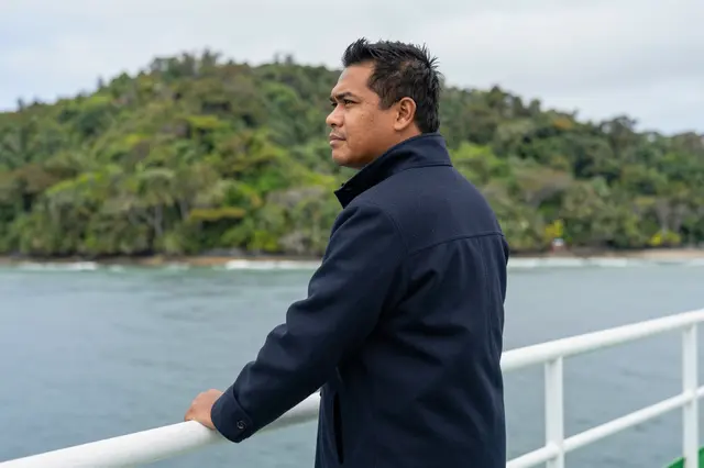 A Pacific Islander man in a navy coat stands leaning on a ferry rail overlooking forested coastal islands.