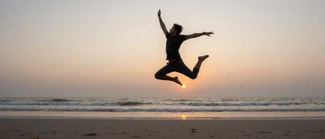 Silhouette of a man mid-leap in the air above a quiet beach as the sun sets over the ocean