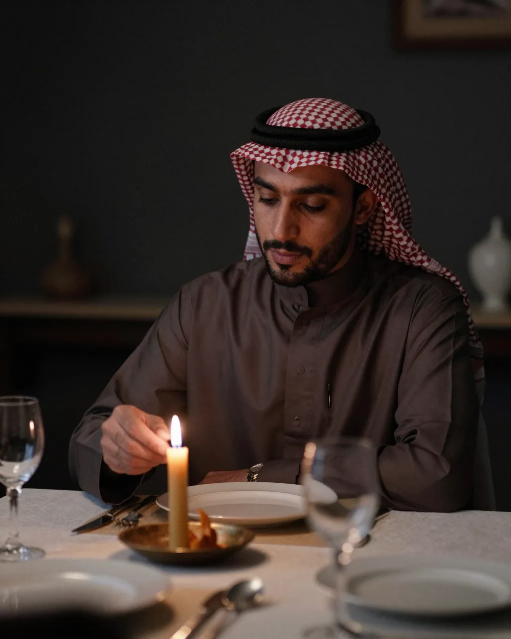 Middle Eastern man in traditional attire lights a candle at a dimly lit dinner table.