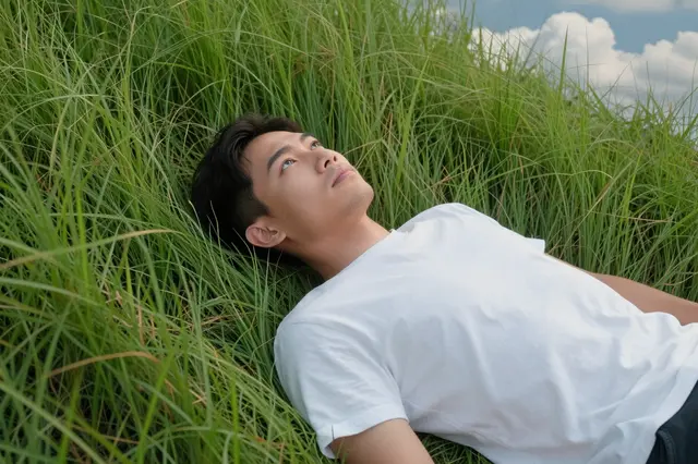 A Southeast Asian man in his late twenties lies back in tall grass looking up at the sky.