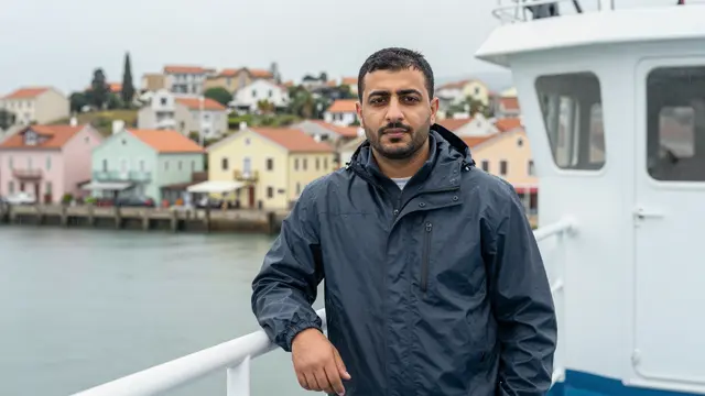 A man in a dark rain jacket stands on a ferry deck overlooking a pastel waterfront town in light rain