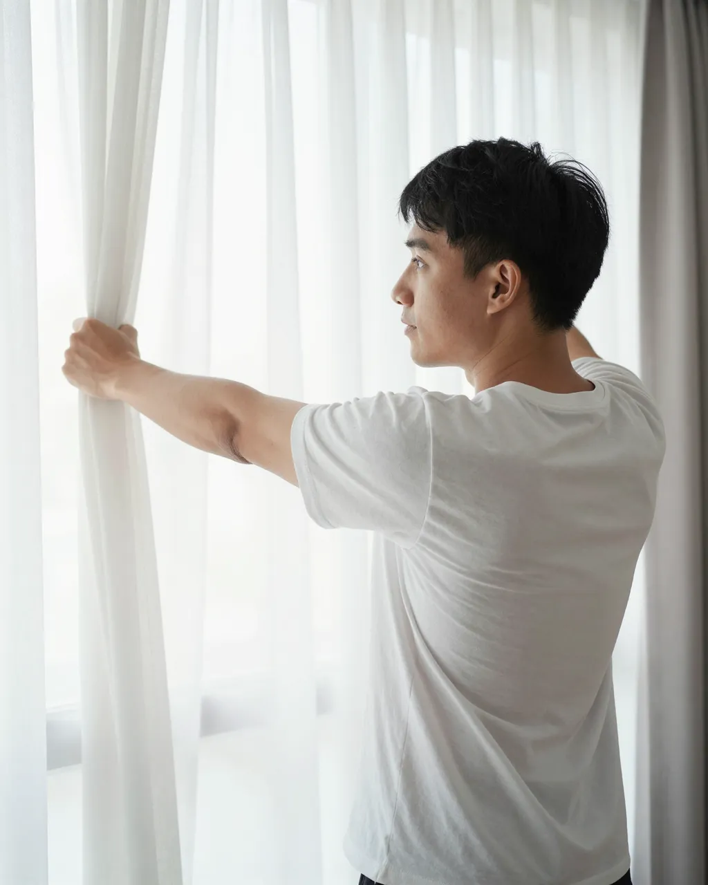 A Southeast Asian man in a plain white t-shirt opens sheer curtains to let in bright morning light.