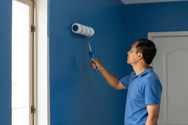 Middle aged Southeast Asian man paints a blue hallway wall with a paint roller next to a sunlit window.