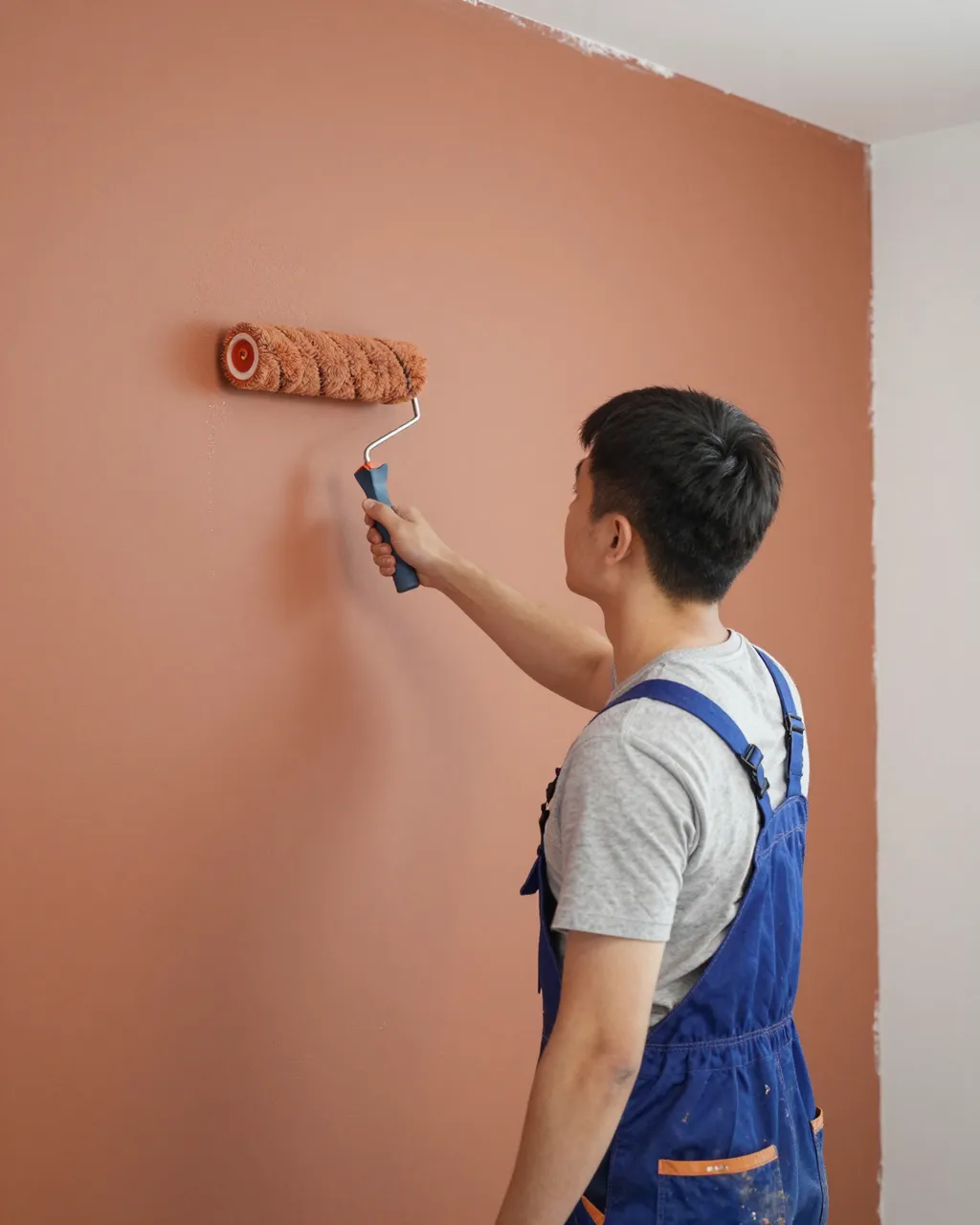 Side view of an East Asian man using a paint roller to apply terracotta paint to an interior wall