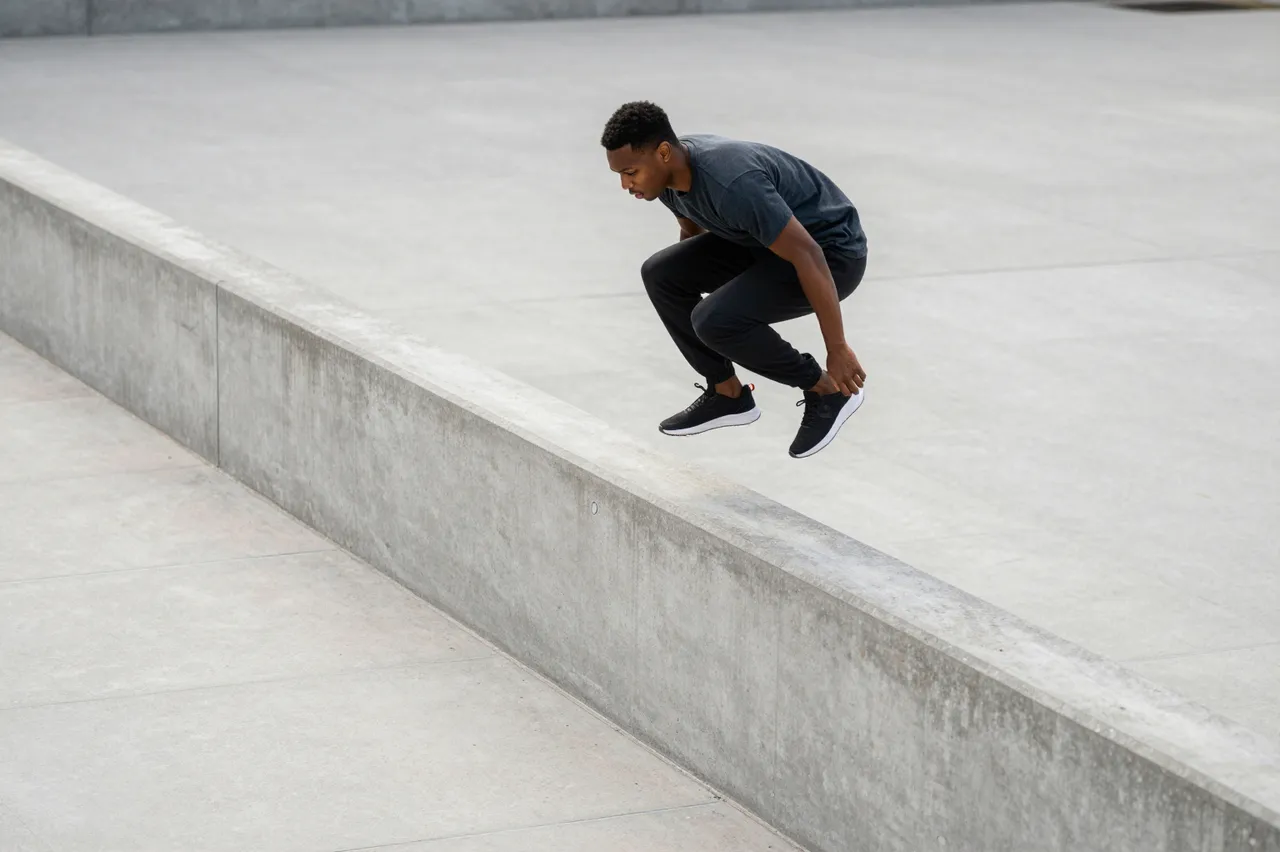 A young Black man mid-jump vaults over a low concrete wall in an empty paved plaza