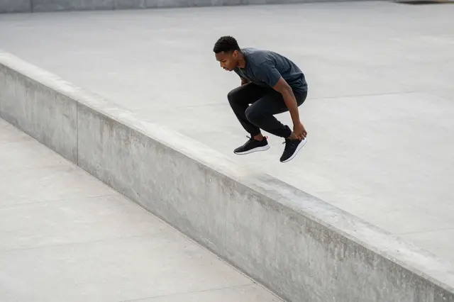 A young Black man mid-jump vaults over a low concrete wall in an empty paved plaza