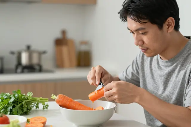 A Southeast Asian man peels a carrot over a white bowl at his bright kitchen table
