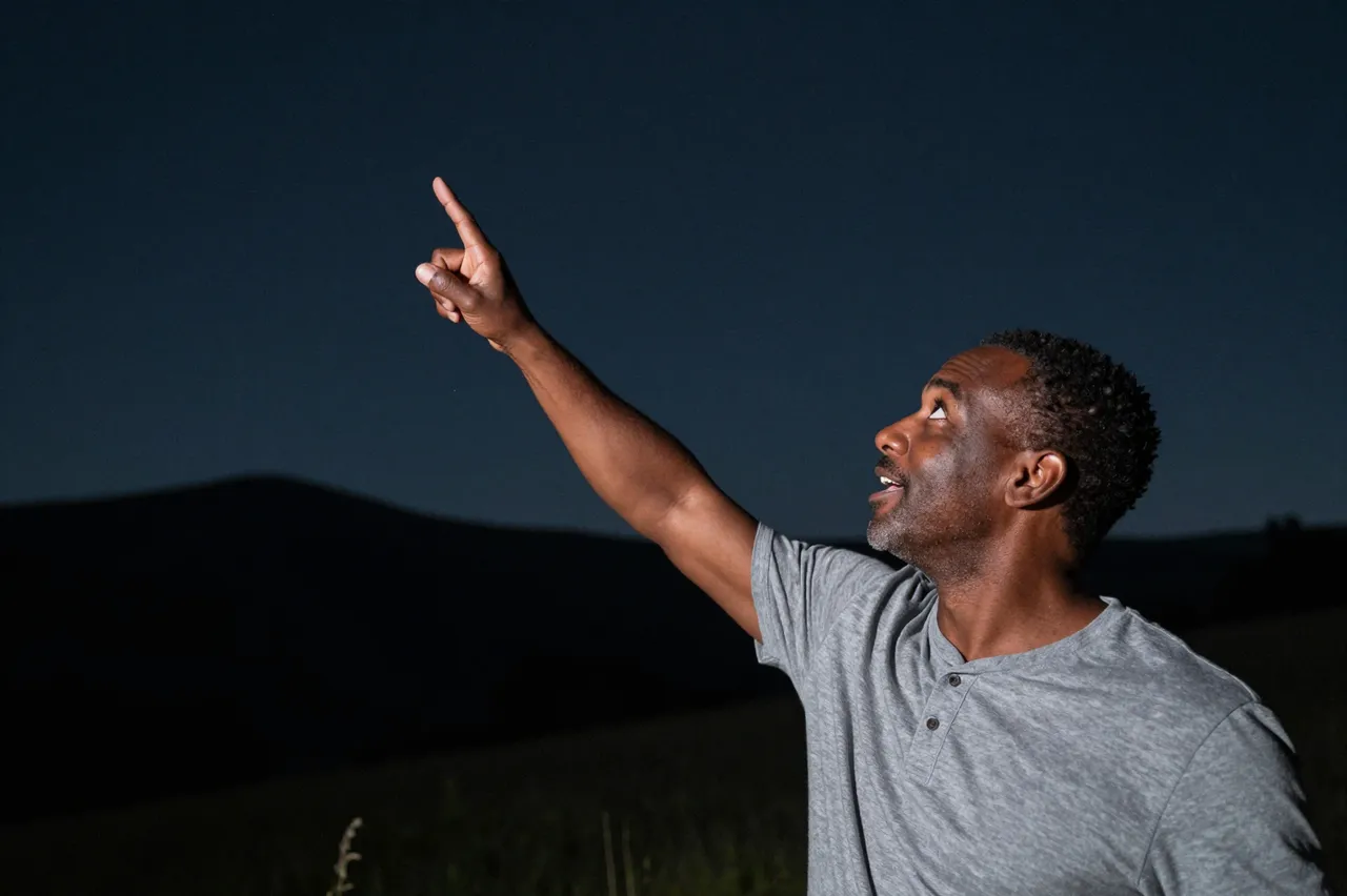 An excited mixed-race man in his forties points upward at the dark night sky above a distant hillside.