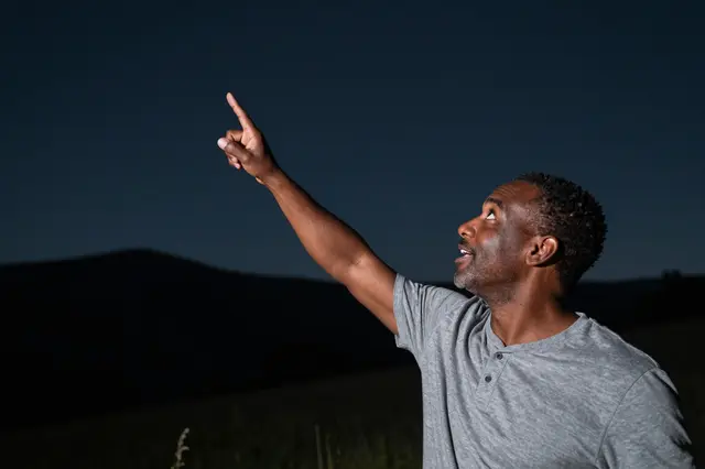 An excited mixed-race man in his forties points upward at the dark night sky above a distant hillside.
