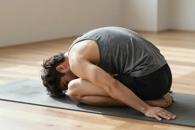 A man in a gray tank top holds a seated forward fold pose on a yoga mat in a sunlit studio