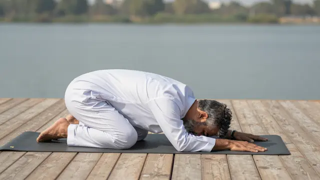 Older man in white clothing holds a forward fold yoga pose on a mat on a wooden lake deck.