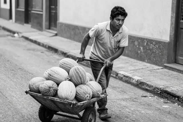 Black and white photo of a Latino man in his thirties pushing a handcart full of melons up a sloped residential street.
