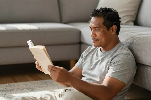 Pacific Islander man sits on the floor leaning against a sofa while reading a book in soft morning sunlight