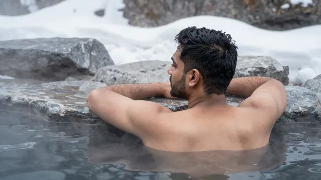 A South Asian man rests in a steaming outdoor thermal pool surrounded by snow covered rocks