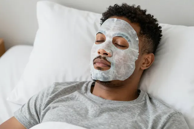 A mixed-race man relaxes in bed wearing a facial sheet mask and gray t-shirt with closed eyes.