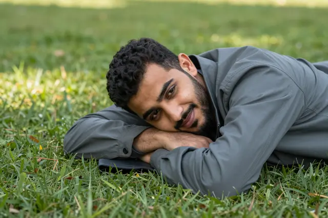 A young Middle Eastern man rests on grass with his head on folded arms, smiling gently at the camera.