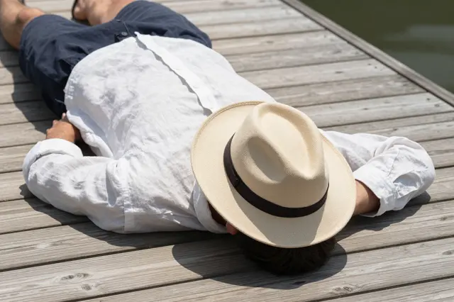 A man in a white linen shirt lies face down on a wooden dock with a straw hat covering his face