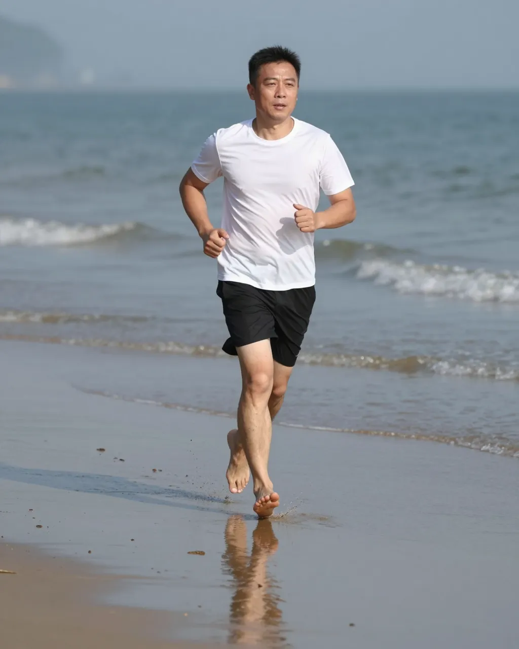 East Asian man in his forties runs barefoot along the wet sandy edge of a beach at midday, his reflection visible in the