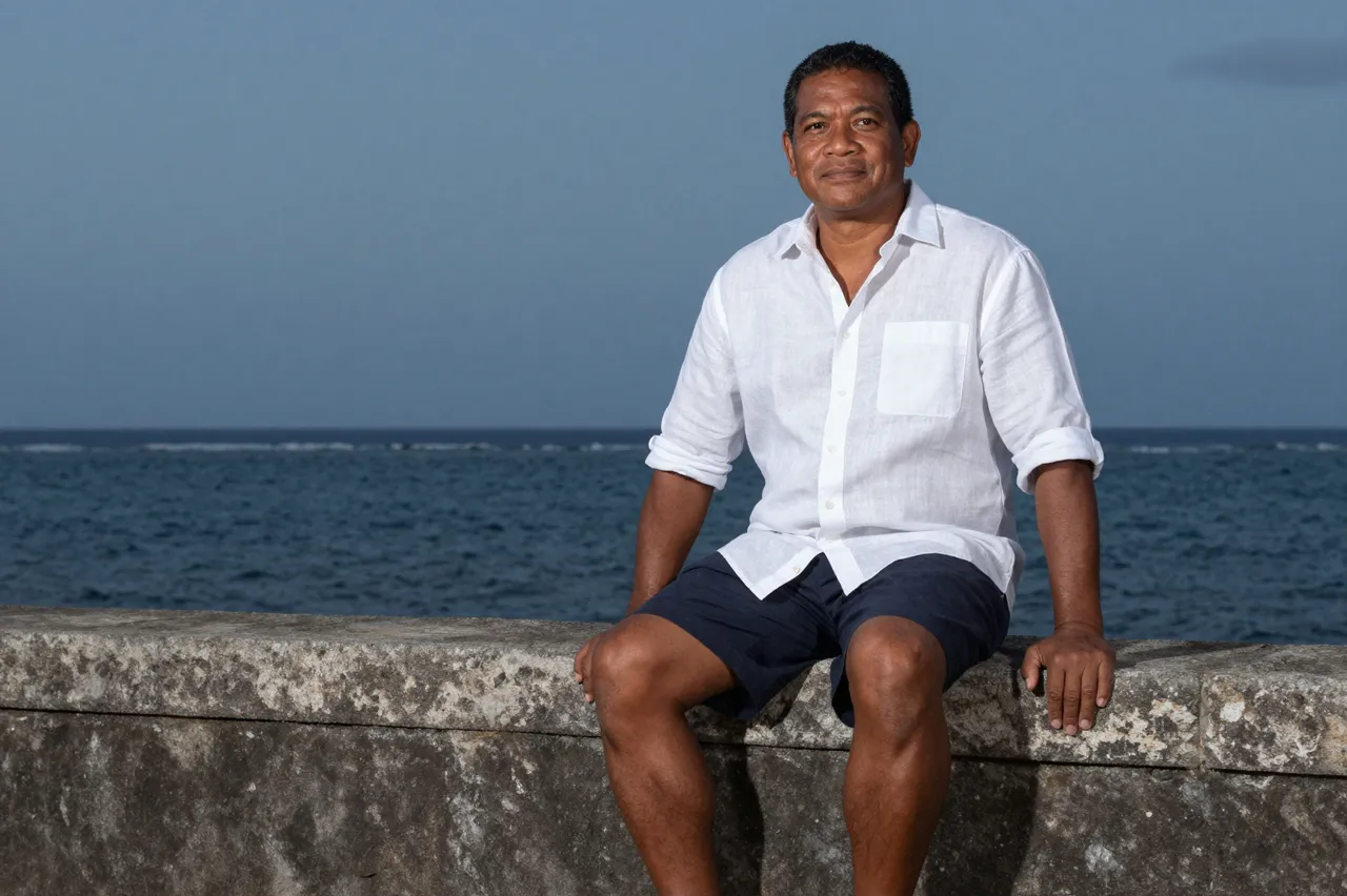Pacific Islander man in a white linen shirt sits on a stone sea wall overlooking the calm ocean at dusk