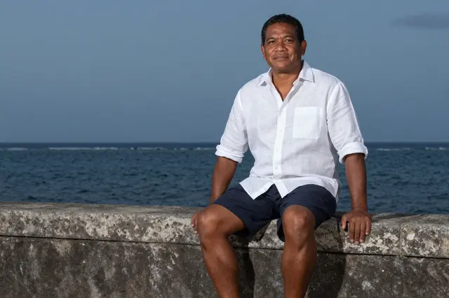 Pacific Islander man in a white linen shirt sits on a stone sea wall overlooking the calm ocean at dusk