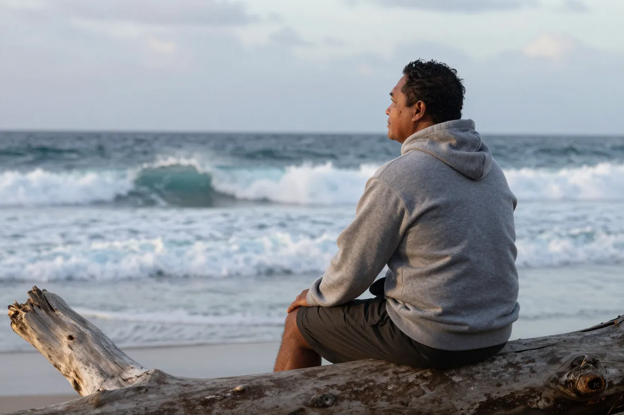 Side view of a Pacific Islander man sitting on driftwood watching ocean waves roll in at dusk
