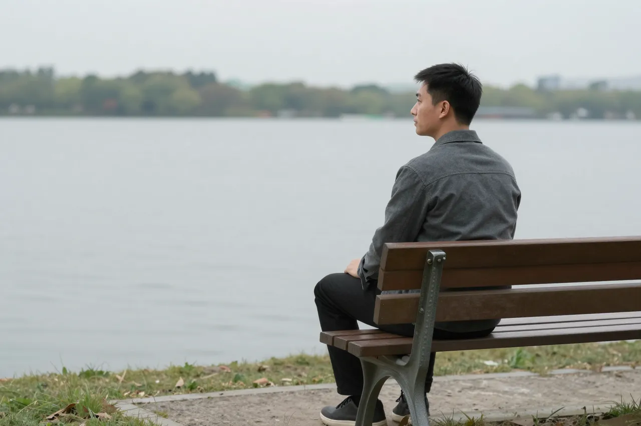A man in his thirties sits on a wooden lakeside bench in side view, looking out over calm gray water on an overcast day.