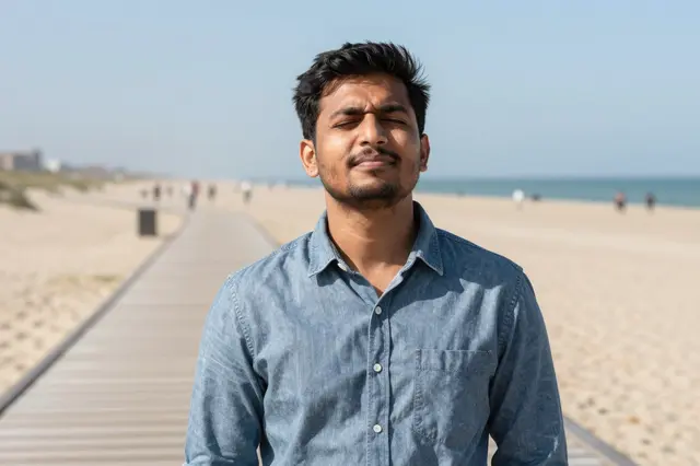 South Asian man in a denim shirt squints into bright sun on a beach boardwalk