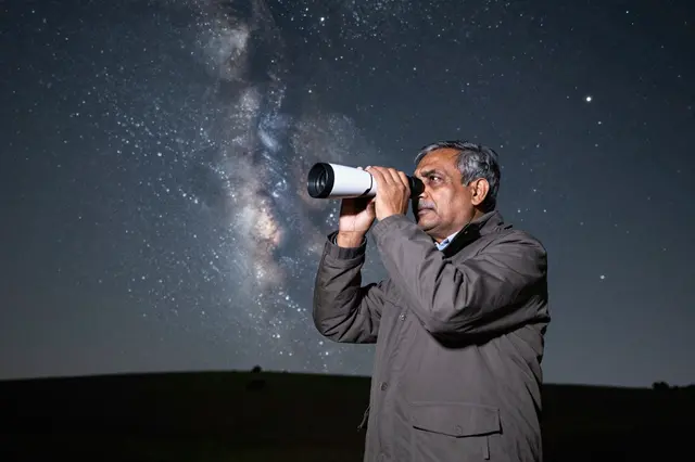 Older South Asian man looks through a small white telescope beneath the bright Milky Way from a hilltop at night