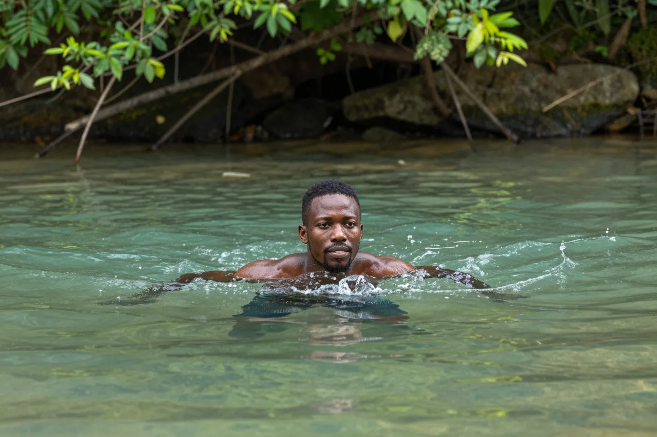 A Black man in his late twenties swims with his head above clear green river water beneath overhanging trees.