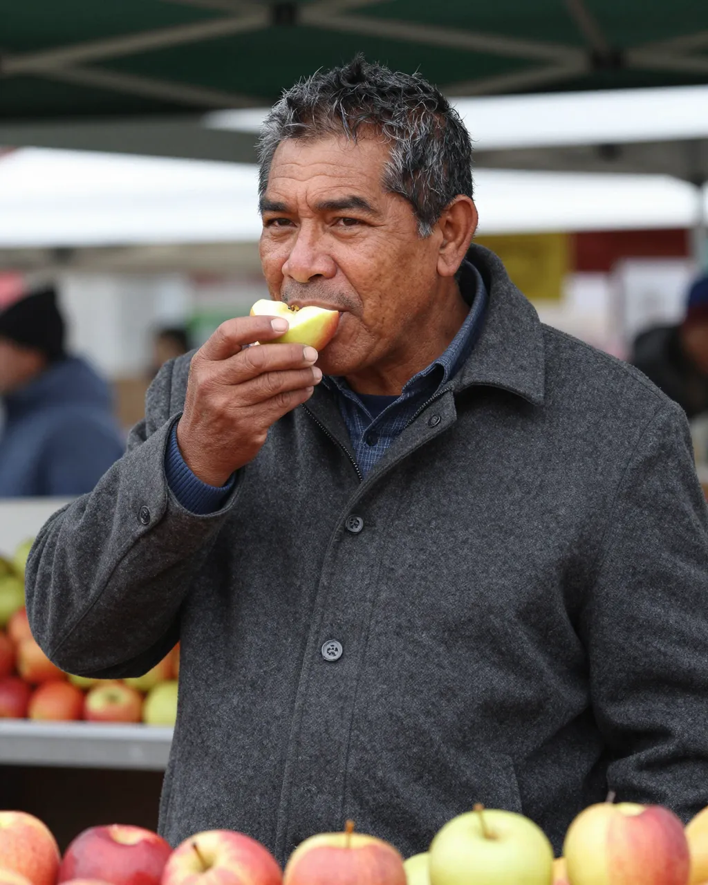 A middle aged man in a gray wool coat takes a bite of fresh apple at an outdoor winter market stall.
