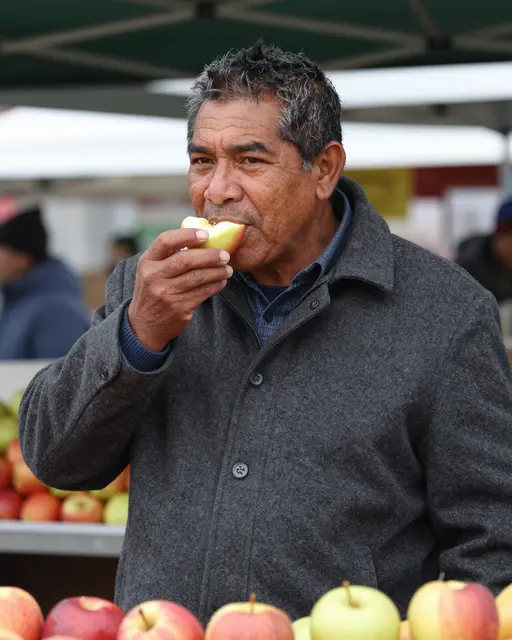 A middle aged man in a gray wool coat takes a bite of fresh apple at an outdoor winter market stall.