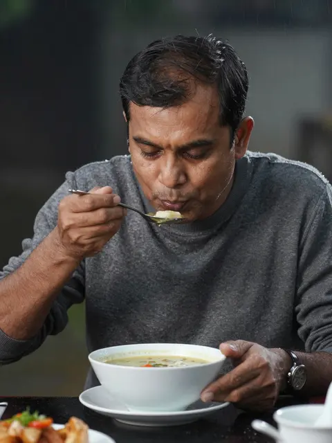 South Asian man in a gray sweater blows on a spoonful of soup from a large white bowl outdoors in rain.