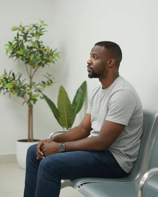 Side view of a Black man in his forties sitting alone on a clinic waiting room chair next to potted plants