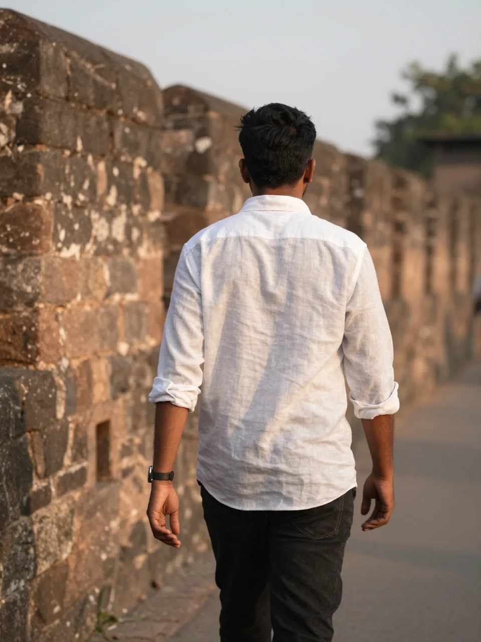 A South Asian man viewed from behind walks beside an old stone fort wall in warm sunset light.