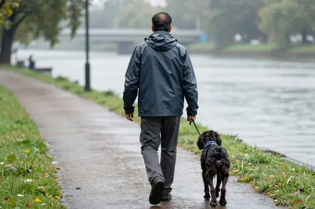 A man in a rain jacket walks a spaniel on a riverside path during light rain, viewed from behind.